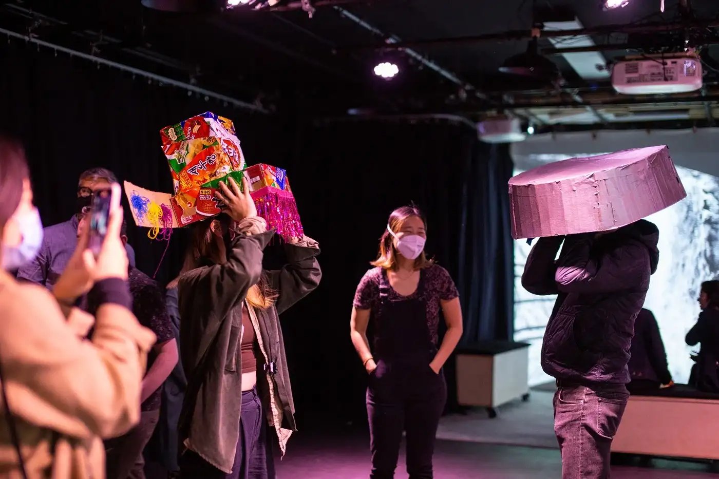 A group faces each other in the dark CultureHub studio, one wearing a cardboard washbasin on their head, while another wears a multicolored Korean crown covered with snack wrappers. Ann looks on from the back, wearing a mask.